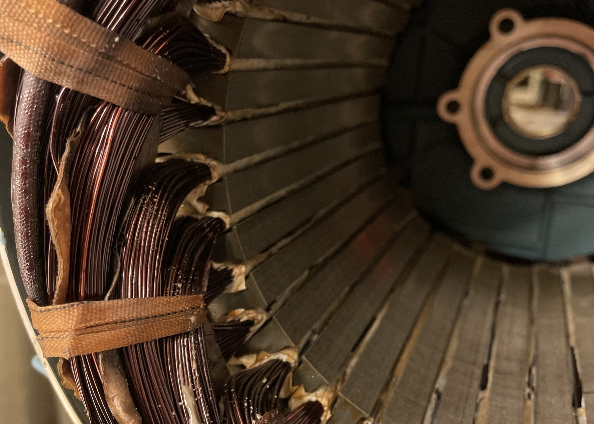 Color photograph looking into an opened electric motor stator: dark copper wire windings bound with fabric tape on the left, pale insulation paper protruding from slots in a laminated steel core that recedes toward the right, and a round end bell with a bearing seat at the far right.